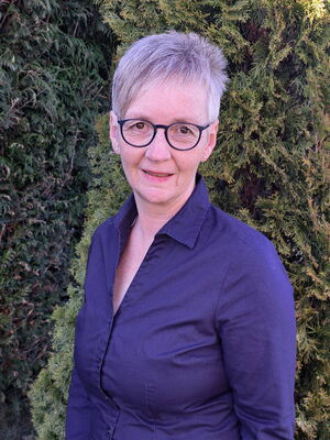 Eine Person mit Brille und grauen Haaren steht vor grünen Hecken, trägt ein blaues Hemd.