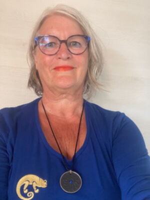 Es ist eine Person mit Brille, einem blauen Oberteil mit einem goldenen Motiv und einer Halskette abgebildet.
