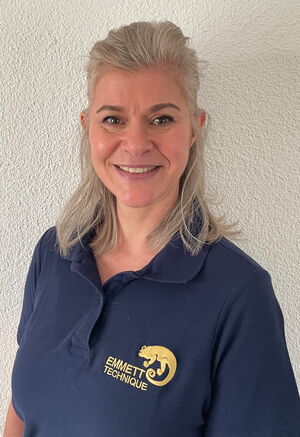 Eine lächelnde Person in einem blauen "Emmett Technique"-Polohemd vor einer weißen Wand.
