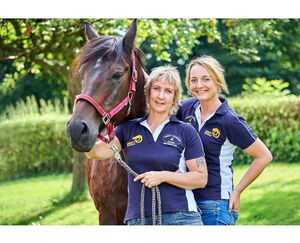 Zwei Personen in Polohemden mit einem Pferd im Freien lächelnd.