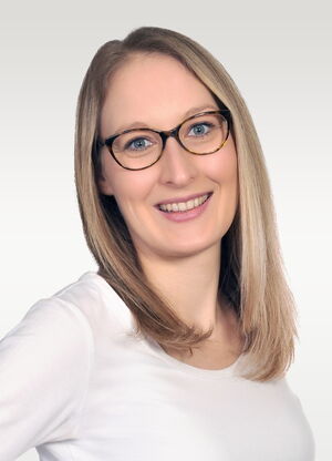Es ist eine lächelnde Person mit Brille und hellbraunem Haar in einem weißen Oberteil vor neutralem Hintergrund.
