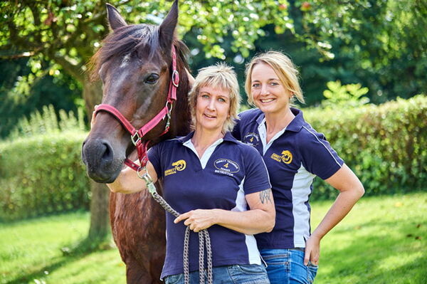 Zwei Personen posieren mit einem Pferd in einem Garten, tragen passende blaue Poloshirts.