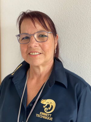 Es ist eine Frau in dunkelblauer Kleidung mit "EMMETT TECHNIQUE"-Aufschrift, mit Brille und lächelnd vor einer weißen Wand.