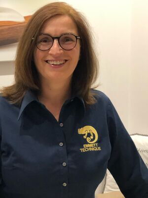 Es ist eine Frau mit Brille in einem Hemd mit dem Logo "Emmett Technique", die lächelt.
