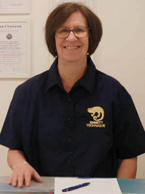 Eine Person lächelt in Arbeitskleidung mit einem goldenen Logo darauf und es hängen Zertifikate an der Wand.