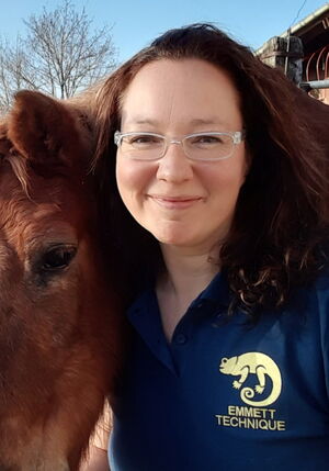 Eine Frau mit Brille und einem Pferd. Sie trägt ein Hemd mit "EMMETT TECHNIQUE"-Logo. Im Hintergrund sind Bäume.