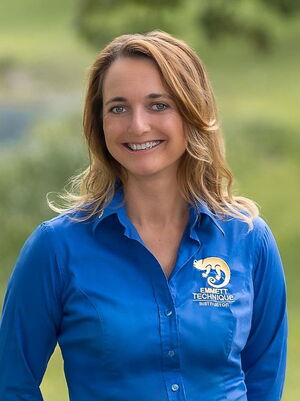 Es zeigt eine Frau in einem blauen Hemd mit "Emmett Technique Instructor"-Logo, lächelnd im Freien.
