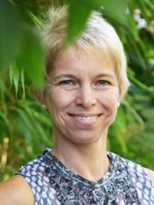 Es zeigt eine lächelnde Person mit kurzem, blondem Haar vor einem Hintergrund mit grünen Blättern.