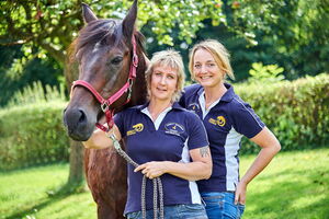 Zwei Personen posieren mit einem Pferd in einem Garten, tragen passende blaue Poloshirts.