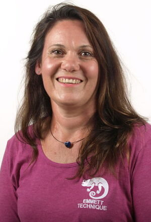 Es ist eine Frau in einem lila Shirt mit der Aufschrift "Emmett Technique" und einem Anhänger. Sie lächelt.