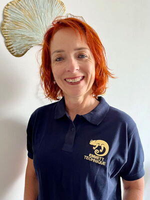 Eine lächelnde Person in einem dunkelblauen Emmett Technique T-Shirt steht vor einer weißen Wand mit Deko.