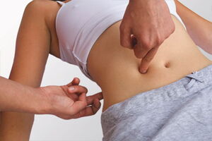 Eine Person zeigt auf den Bauch einer anderen Person, die ein weißes Oberteil und graue Hose trägt.