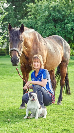 Eine Frau hockt mit ihrem Hund vor einem großen Pferd auf einer grünen Wiese, umgeben von Bäumen.