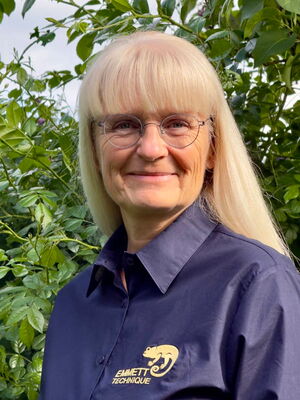 Es zeigt eine Person mit Brille und blondem Haar, die ein Hemd mit "Emmett Technique"-Logo trägt, vor Pflanzen.