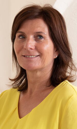 Es ist eine Frau mit schulterlangem braunem Haar, die ein gelbes Oberteil trägt und lächelt.