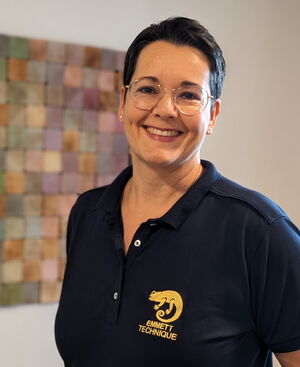 Eine lächelnde Person trägt ein dunkelblaues Polo mit "EMMETT TECHNIQUE"-Logo, vor einem bunten Quadratmuster.