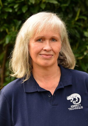 Eine Frau mit blonden Haaren trägt ein dunkelblaues Polohemd mit einem Logo "EMMETT TECHNIQUE" und steht vor Bäumen.
