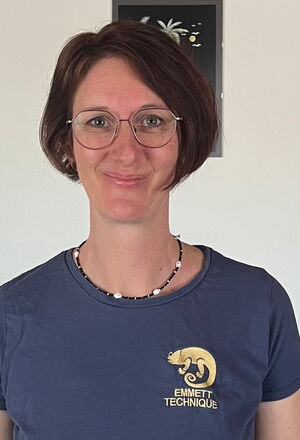 Eine Frau mit Brille und Kette trägt ein T-Shirt mit "EMMETT TECHNIQUE"-Logo und einem Chamäleon darauf.