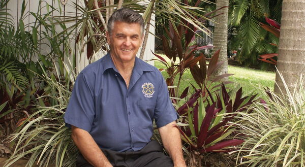 Ein Mann in blauer Uniform sitzt lächelnd draußen, umgeben von tropischen Pflanzen und Bäumen.