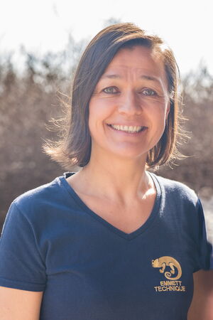 I see a person smiling, wearing a navy blue shirt with "EMMETT TECHNIQUE" and a chameleon logo printed in yellow. The background appears to be outdoors with blurred foliage.