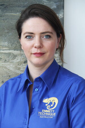 Person in blauem Hemd mit "Emmett Technique Instructor"-Logo, vor einer Wand stehend.