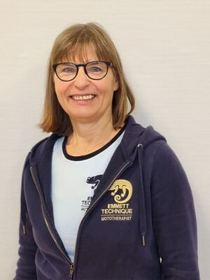 Eine Frau mit Brille lächelt. Sie trägt eine Jacke mit dem Logo "Emmett Technique" und der Aufschrift "Mototherapist".