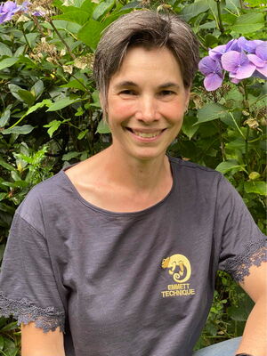 Eine Person lächelt in einem Garten, trägt ein T-Shirt mit "Emmett Technique" und steht vor lila Blumen.